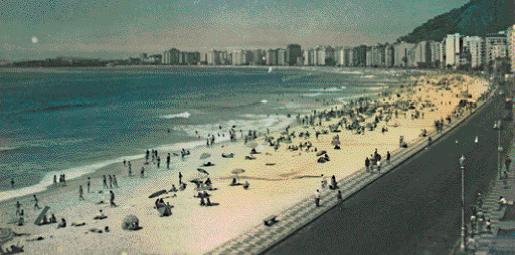 Imagem: Fotografia em preto e branco. Várias pessoas na orla da praia. À direita, automóveis na rua e em seguida, prédios. Fim da imagem.