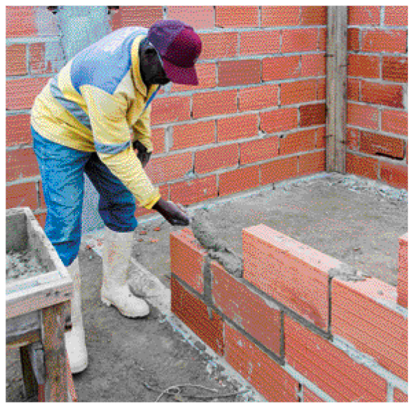 Imagem: Fotografia. Um homem está colocando cimento em uma parede de tijolos.  Fim da imagem.