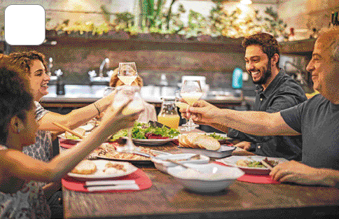 Imagem: Fotografia. Um casal, um senhor e uma menina estão sentados em volta de uma mesa com comida e brindando com copos.   Fim da imagem.