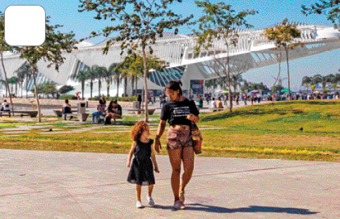 Imagem: Fotografia. Uma mulher e uma criança estão andando em um parque. Ao fundo, árvores, pessoas e uma construção grande.   Fim da imagem.