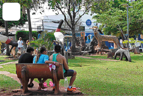 Imagem: Fotografia. Três adultos sentados e observando crianças brincando em um parque com estátuas de animais.   Fim da imagem.