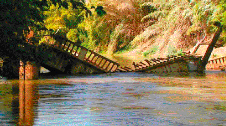 Imagem: Fotografia. Uma ponte quebrada sobre um rio. Ao fundo, plantas.  Fim da imagem.