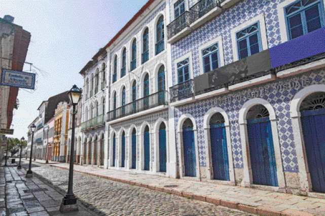 Imagem: Fotografia. Construções lado a lado com paredes de azulejo azul. Ao lado, rua de pedras e postes.   Fim da imagem.