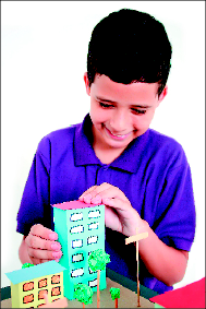 Imagem: Fotografia. Um menino está sorrindo e colando um prédio de papel em uma maquete.   Fim da imagem.