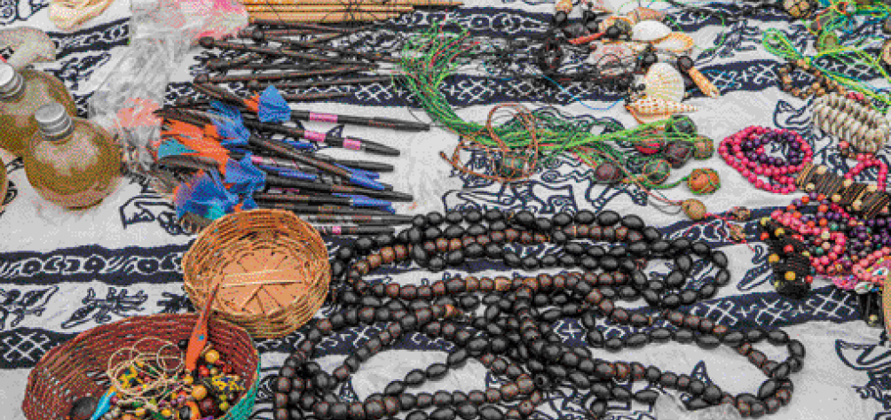 Imagem: Fotografia. Vários objetos sobre um tecido branco e azul: canetas com penas coloridas, potes, cestos com miçangas, colares e pulseiras. Fim da imagem.