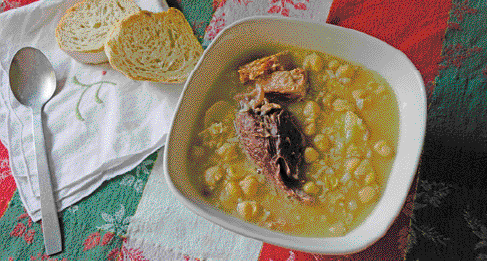 Imagem: Fotografia. Uma tigela branca e dentro há uma sopa amarela com grãos e pedaços de carne. Ao lado há dois pedaços de pão e uma colher. Fim da imagem.