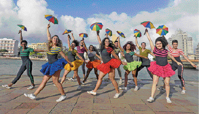 Imagem: Fotografia. Pessoas estão segurando guarda-chuvas coloridos com as mãos levantadas e sorrindo. Ao fundo, prédios.   Fim da imagem.