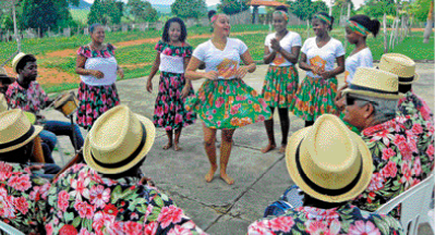 Imagem: Fotografia. Homens e mulheres com roupas floridas estão em roda e cantando. No centro, uma mulher está dançando. Ao fundo, árvores.  Fim da imagem.