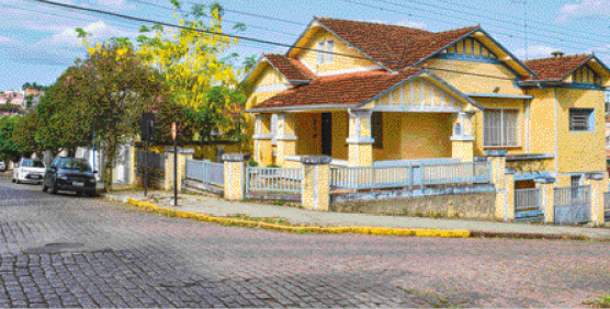 Imagem: Fotografia. Uma casa grande com paredes amarelas e detalhes azuis. Ao lado há árvores na calçada e em volta, uma rua de paralelepípedos.  Fim da imagem.