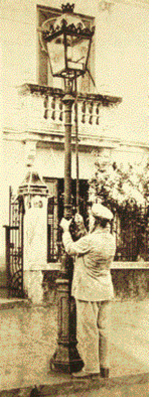 Imagem: Fotografia em sépia. Um homem com boina e terno está segurando uma haste comprida dentro de uma lamparina de um poste. Ao fundo, uma construção.   Fim da imagem.