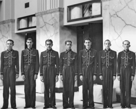 Imagem: Fotografia em preto e branco. Sete homens com camisa de manga comprida, calça e sapatos estão lado a lado e olhando para frente. Ao fundo, salão grande com duas portas.   Fim da imagem.