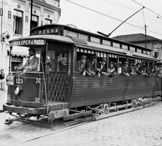 Imagem: Fotografia em preto e branco. Várias pessoas sentadas em um bonde, que está em cima de um trilho. Na parte da frente, um homem está em pé e conduzindo o bonde. Ao fundo, construções.   Fim da imagem.