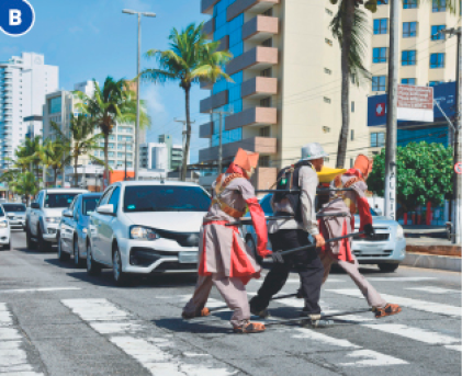 Imagem: Fotografia B. Uma pessoa está segurando dois suportes na lateral do corpo e andando sobre uma faixa de pedestres. Nas pontas dos suportes há dois bonecos pendurados. Ao fundo, carros enfileirados e prédios.  Fim da imagem.