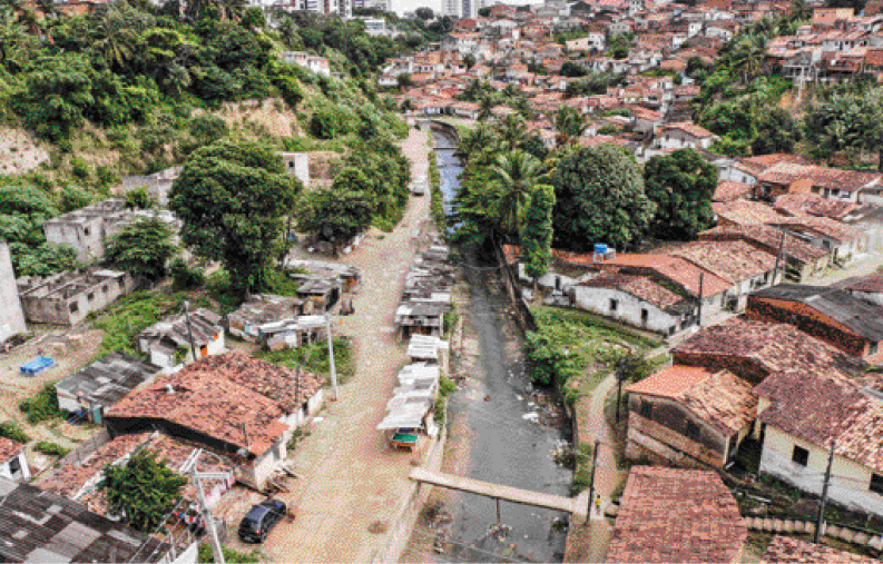 Imagem: Fotografia. Vista aérea de um rio com água escura e lixo nas margens. Nas laterais há casas, árvores e postes.  Fim da imagem.