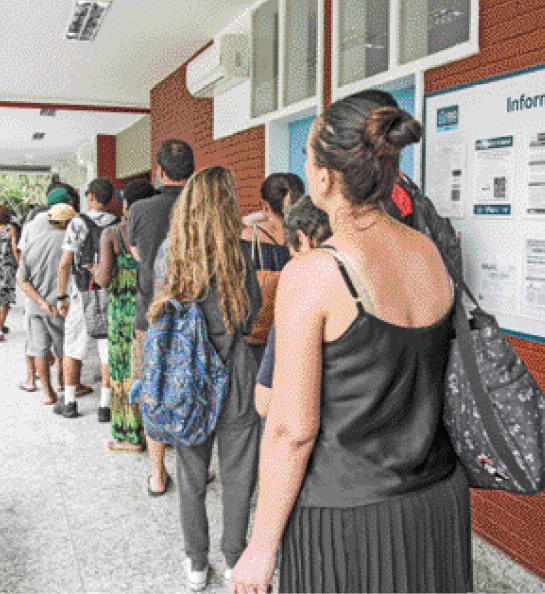 Imagem: Fotografia. Várias pessoas enfileiradas na frente de uma porta.   Fim da imagem.