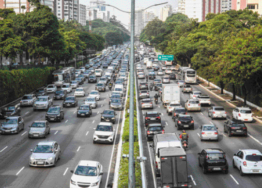 Imagem: Fotografia. Vários automóveis enfileirados e duas avenidas. Nas laterais há árvores e construções.   Fim da imagem.