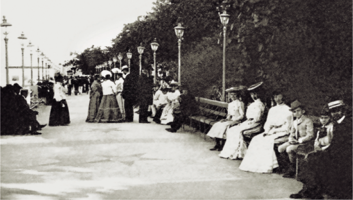 Imagem: Fotografia em preto e branco. No centro, homens com ternos e mulheres com vestidos estão em pé e conversando. Nas laterais há homens e mulheres sentados em bancos e atrás deles há árvores e postes. Fim da imagem.