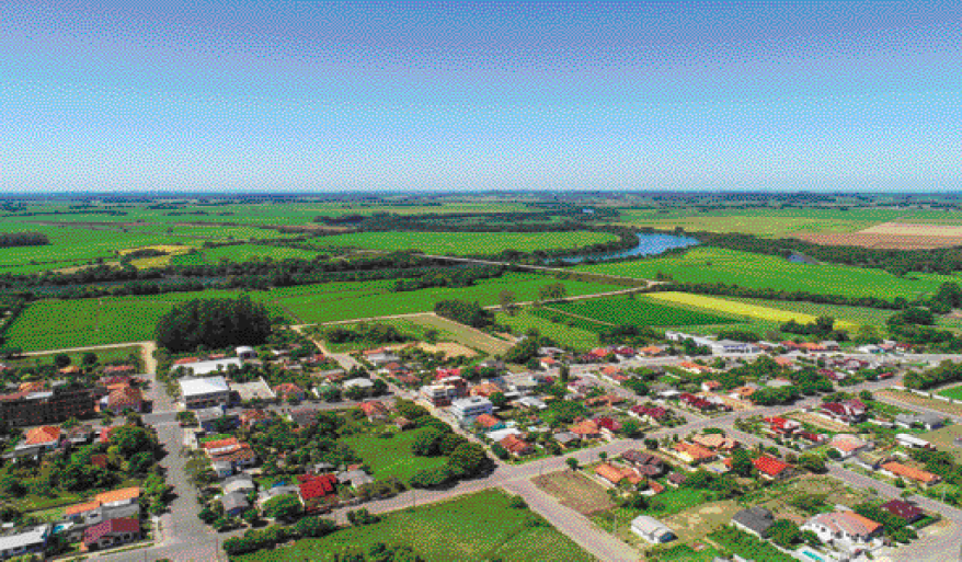 Imagem: Fotografia. Vista aérea de uma cidade. Na parte inferior há casas, árvores e ruas asfaltadas. Na parte superior, campos com plantações, árvores e um rio sinuoso. Fim da imagem.