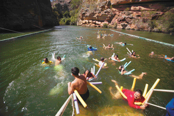 Imagem: Fotografia. Várias pessoas nadando em um rio. Ao fundo, morros e plantas.  Fim da imagem.