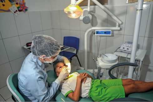 Imagem: Fotografia. Uma dentista está sentada e com as mãos dentro da boca de uma criança, que está sentada na sua frente. Fim da imagem.