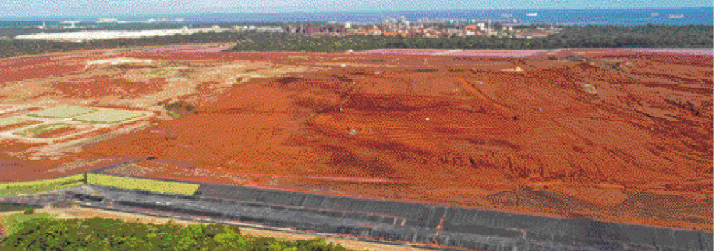 Imagem: Fotografia. Vista aérea de um terreno plano e marrom. Ao fundo, árvores e uma cidade. Fim da imagem.