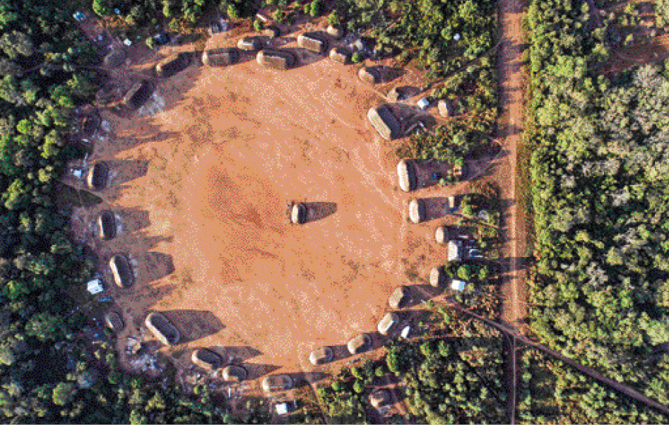 Imagem: Fotografia. Vista aérea de ocas formando um círculo sobre terreno plano. Em volta há muitas árvores e ao lado, uma estrada de terra. Fim da imagem.