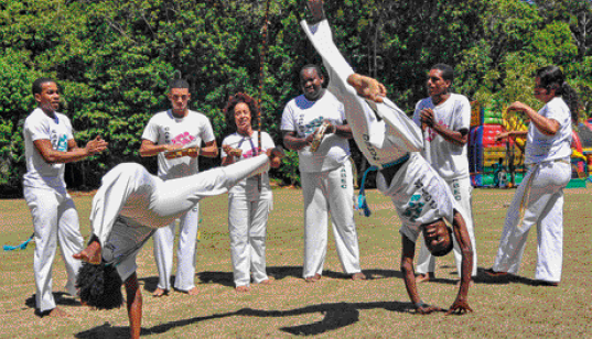 Imagem: Fotografia. Duas pessoas dançando capoeira. Atrás há pessoas tocando instrumentos musicais.  Fim da imagem.