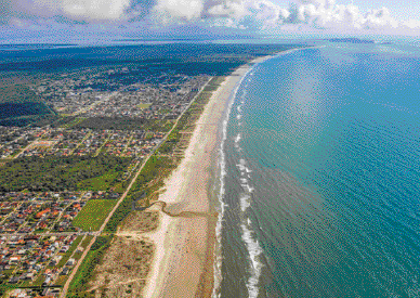 Imagem: Fotografia. Vista aérea do mar e ao lado, casas, árvores e ruas. Fim da imagem.