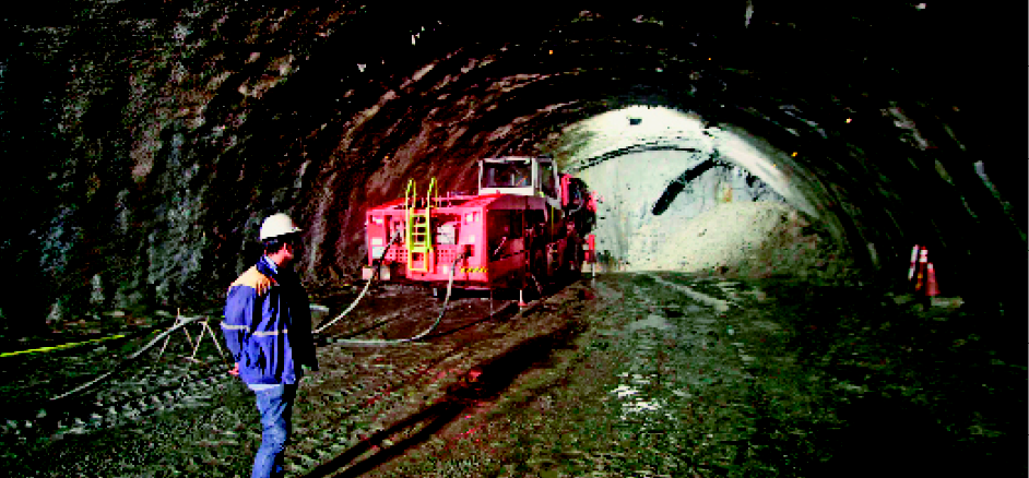 Imagem: Fotografia. Um homem com capacete de proteção e uniforme azul observa um caminhão vermelho dentro de um túnel com terra em volta. Fim da imagem.