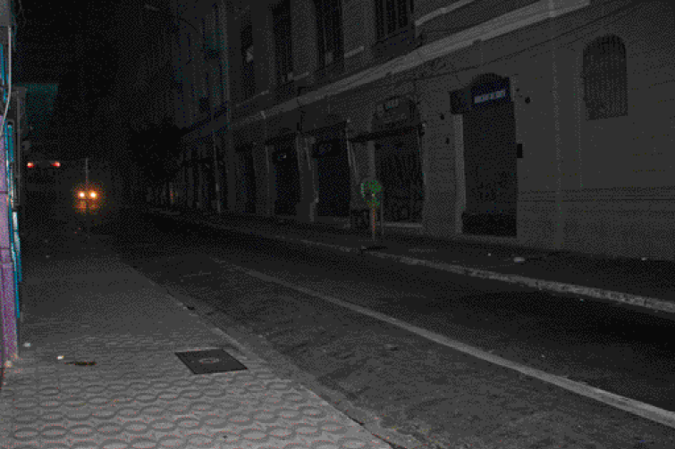 Imagem: Fotografia. À esquerda, rua asfaltada e escura. À direita, sombra de prédios. Ao fundo, faróis de um carro acesso. Fim da imagem.