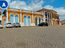 Imagem: Fotografia A. Carros parados sobre uma rua de paralelepípedos e ao lado de construções coloridas.  Fim da imagem.