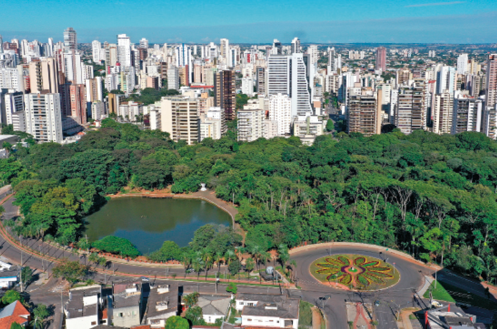 Imagem: Fotografia. Vista aérea de um parque com um lago grande à esquerda e muitas árvores em volta. Ao redor do parque há ruas asfaltadas, casas e postes. Ao fundo, prédios.  Fim da imagem.