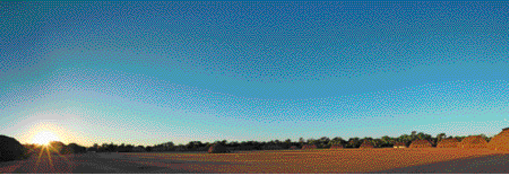 Imagem: Fotografia. Campo extenso com ocas em volta e árvores ao fundo. À esquerda, o sol se pondo no céu em tons de azul.  Fim da imagem.