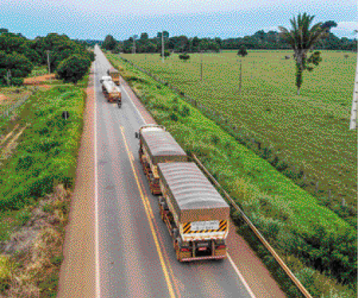 Imagem: Fotografia. Vista aérea de caminhões em uma estrada asfaltada. Nas laterais há plantas e árvores. Fim da imagem.