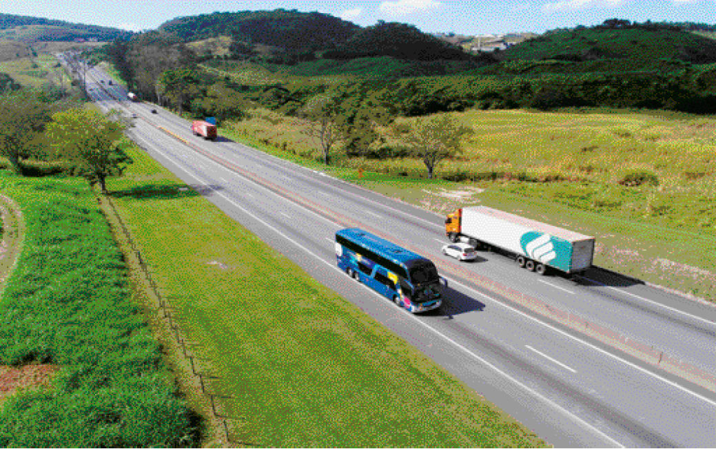 Imagem: Fotografia. Vista aérea de um ônibus em uma estrada asfaltada. Do outro lado, no caminho inverso há um caminhão e um carro. Ao fundo, mais automóveis na estrada. Nas laterais há plantas e árvores e atrás, morros com vegetação. Fim da imagem.