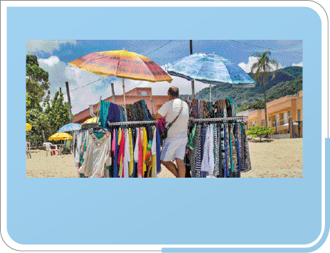 Imagem: Fotografia. Um homem está de costas e na frente dele há várias roupas coloridas penduradas em uma barra de ferro. Acima dele, dois guarda-sóis coloridos e ao fundo, areia da praia, construções e árvores. Fim da imagem.