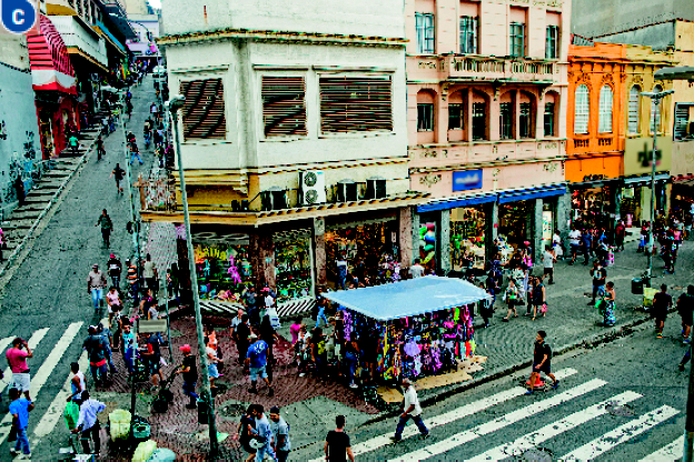 Imagem: Fotografia C. Vista aérea de uma rua cheia de pessoas. No centro há lojas e barracas. Fim da imagem.