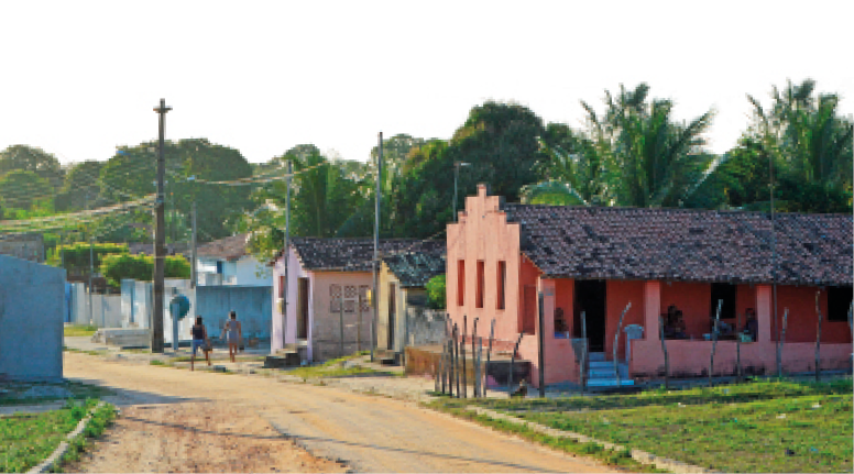 Imagem: Fotografia. Casas ao lado de uma estrada de terra. Ao fundo, árvores. Fim da imagem.