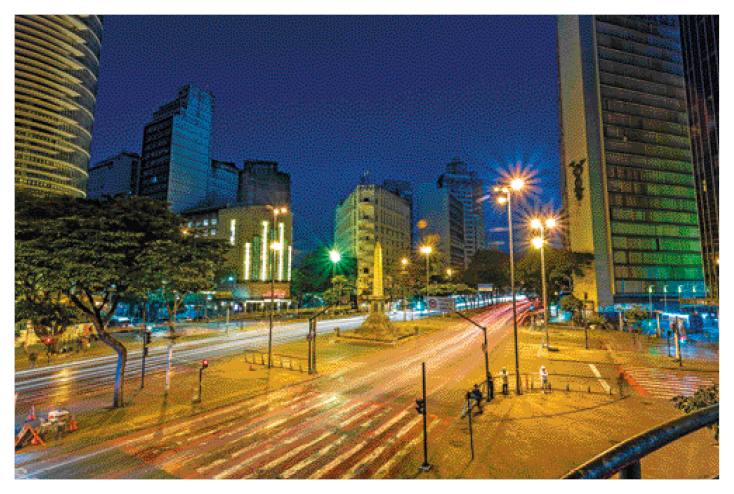 Imagem: Fotografia. Uma avenida e nas laterais há postes acesos. Ao fundo, prédios e árvores. Fim da imagem.