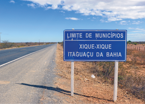 Imagem: Fotografia. À esquerda, estrada asfaltada e à direita, placa azul com a informação: LIMITE DE MUNICÍPIOS – XIQUE-XIQUE – ITAGUAÇU DA BAHIA.  Fim da imagem.