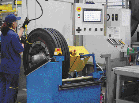 Imagem: Fotografia. Uma mulher com uniforme está manuseando uma máquina. Ao seu lado, um pneu.  Fim da imagem.