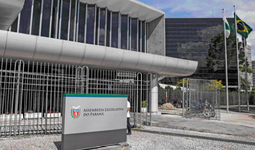 Imagem: Fotografia. Placa com um brasão e a informação: Assembleia Legislativa do Paraná. Atrás há construção alta e cerca em volta.  Fim da imagem.