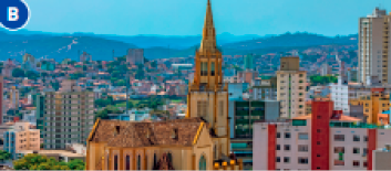 Imagem: Fotografia B. Vista aérea de uma cidade com ruas, prédios, casas e árvores. Ao fundo, morros.  Fim da imagem.