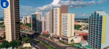 Imagem: Fotografia E. Vista aérea de uma cidade com ruas, prédios, casas e árvores.  Fim da imagem.