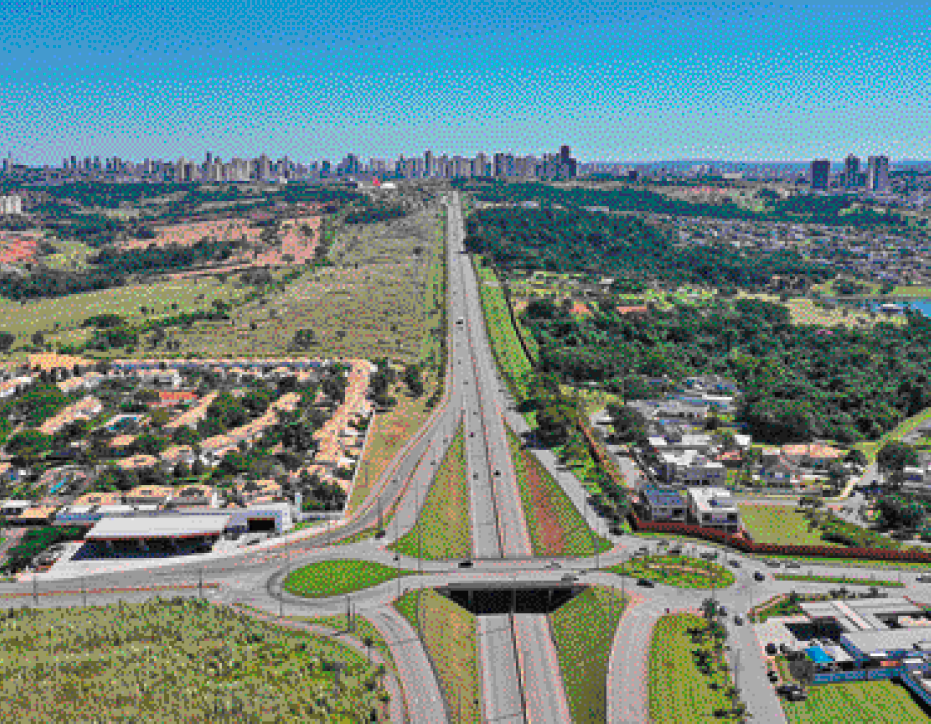 Imagem: Fotografia. Vista aérea de uma estrada asfaltada com rotatórias. Nas laterais há campos verdes, muitas árvores, algumas casas e ruas. Ao fundo, prédios.  Fim da imagem.