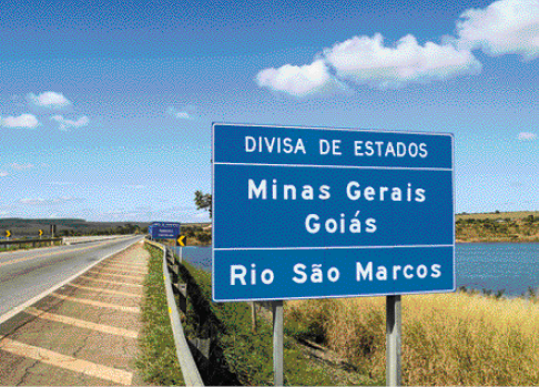 Imagem: Fotografia. À esquerda, estrada asfaltada e à direita, placa azul com a informação: DIVISA DE ESTADOS – MINAS GERAIS – GOIÁS – RIO SÃO MARCOS. Ao fundo, um rio com plantas em volta. Fim da imagem.