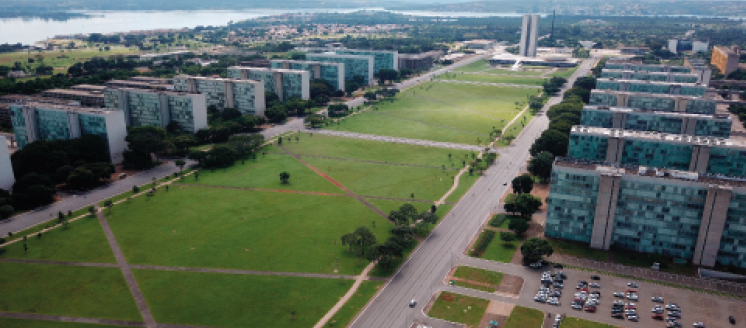 Imagem: Fotografia. Vista aérea de um pátio gramado com árvores. Nas laterais há fileiras de prédios compridos.  Fim da imagem.