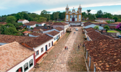 Imagem: Fotografia. Vista aérea de pessoas andando em uma rua de pedras. Nas laterais há casas pequenas com paredes brancas e telhados marrons. Ao fundo, uma igreja e árvores.   Fim da imagem.