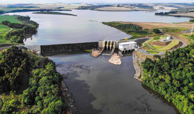 Imagem: Fotografia. Vista aérea de um lago ao lado de uma barragem. Ao redor há vegetação e árvores.   Fim da imagem.