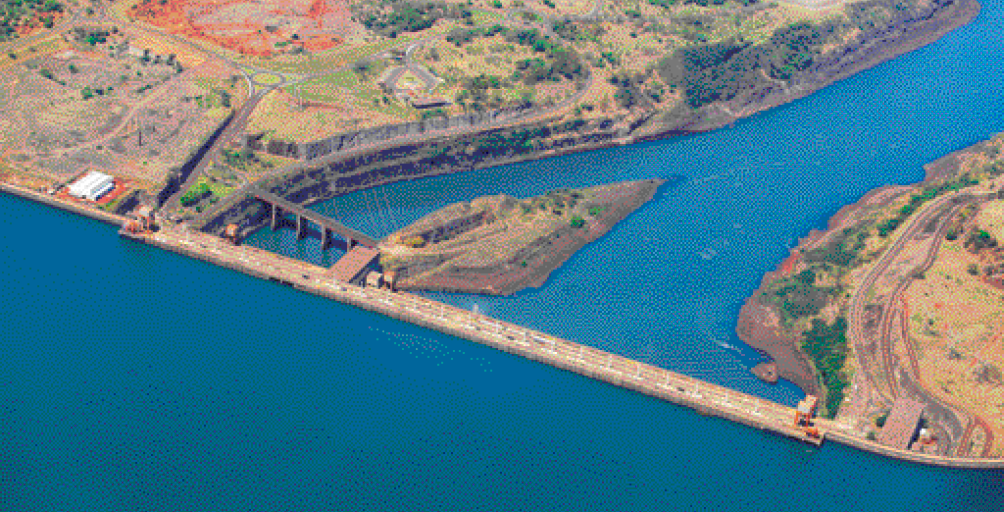 Imagem: Fotografia. Vista aérea de um rio grande com água azul. No centro há uma ponte e nas laterais, estradas, árvores e algumas construções. Fim da imagem.